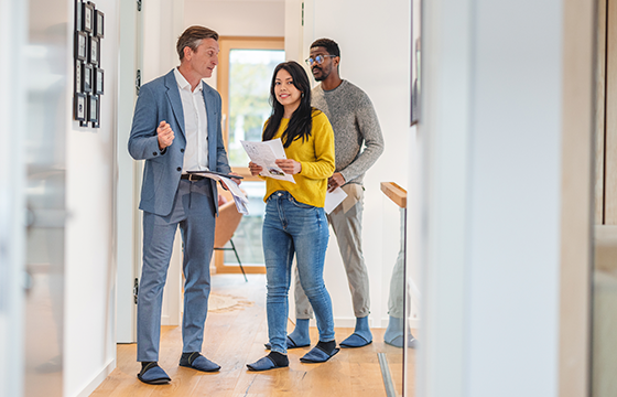 A man and woman walking through a house holding paperwork with a real estate agent in a blue suit.
