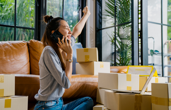 Young Asian woman on cell phone smiling with arm raised in the air surrounded by boxes