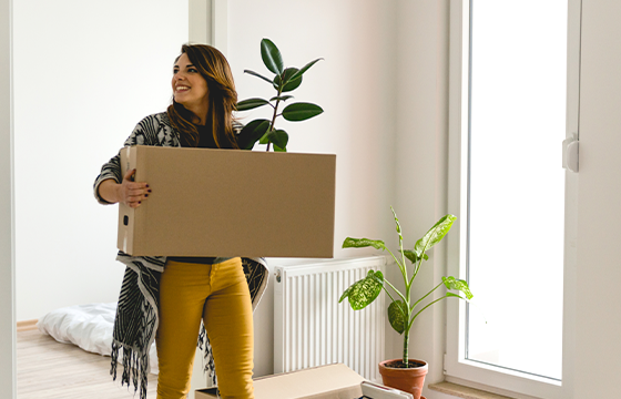 Woman in yellow pants a long black & white cardigan carrying a sealed moving box