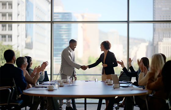 Businesspeople sitting around a conference table and at the head of the table is a man and woman shaking hands
