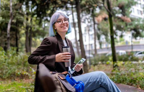 Young intern woman sitting in public park after work and drinking coffee from thermos