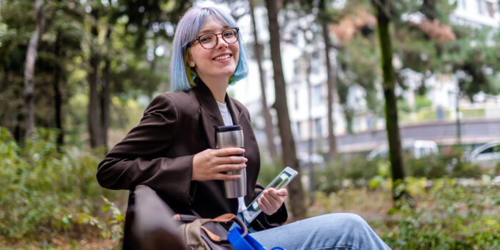 Young intern woman sitting in public park after work and drinking coffee from thermos