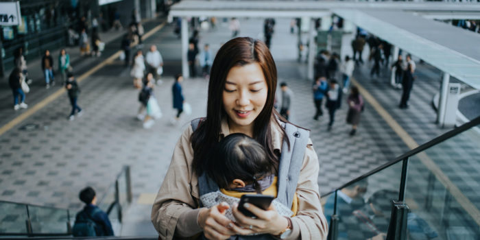 Young Asian mother with little daughter using smartphone while riding on escalator in downtown city, with busy commuters in the background