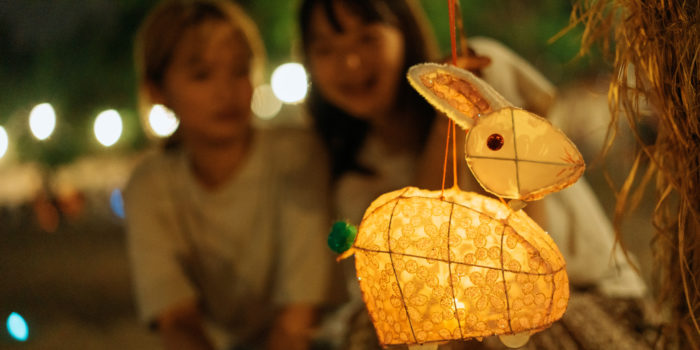A traditional Chinese paper rabbit-shaped lantern hung on a tree as two young happy Asian sisters or female friends celebrate Mid-Autumn Festival
