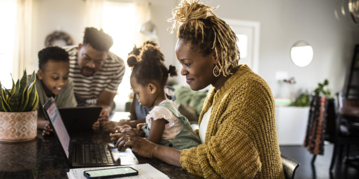 Mother working from home while holding toddler, family in background