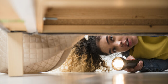 Mixed Race woman looking under bed with flashlight