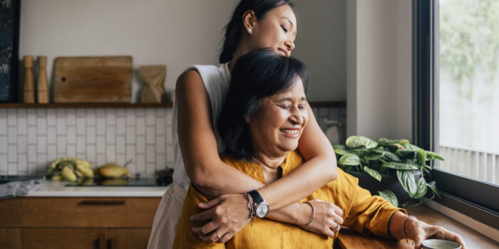 A Happy Beautiful Woman Hugging Her Mother While She Is Sitting In The Kitchen And Drinking Tea