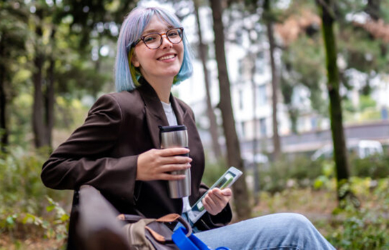 Young woman sitting in public park after work and drinking coffee from thermos