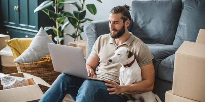 Young men moving in new house. He is happy and using laptop to organize everything. His pet dog is with him.Young men moving in new house. He is happy and using laptop to organize everything. His pet dog is with him.