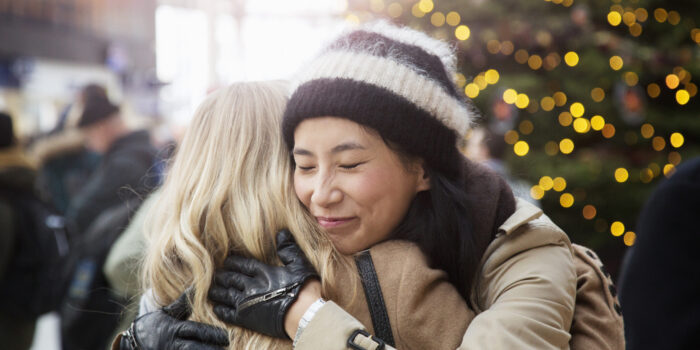 Asian woman embraces friend in railroad station, christmas tree in background.